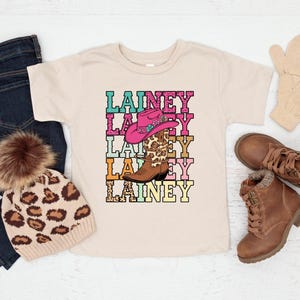 May include: A beige t-shirt with the name "Lainey" stacked vertically in colorful letters. A pink cowboy hat and a brown and leopard print cowboy boot are in the center of the design. A pair of brown boots, a leopard print beanie, and blue jeans are also in the image.