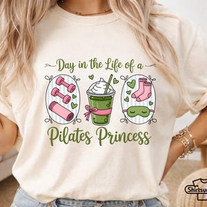 K&ouml;nnte beinhalten: Cremefarbenes T-Shirt mit einem Grafikdesign. Das Design zeigt den Text "Day in the Life of a Pilates Princess" und Illustrationen von Hanteln, einer Yogamatte, einem Getr&auml;nk, Socken und einer Augenmaske in Gr&uuml;n und Rosa.