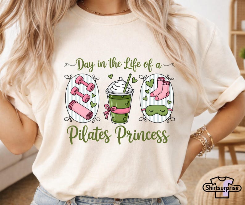 K&ouml;nnte beinhalten: Cremefarbenes T-Shirt mit einem Grafikdesign. Das Design zeigt den Text "Day in the Life of a Pilates Princess" und Illustrationen von Hanteln, einer Yogamatte, einem Getr&auml;nk, Socken und einer Augenmaske in Gr&uuml;n und Rosa.