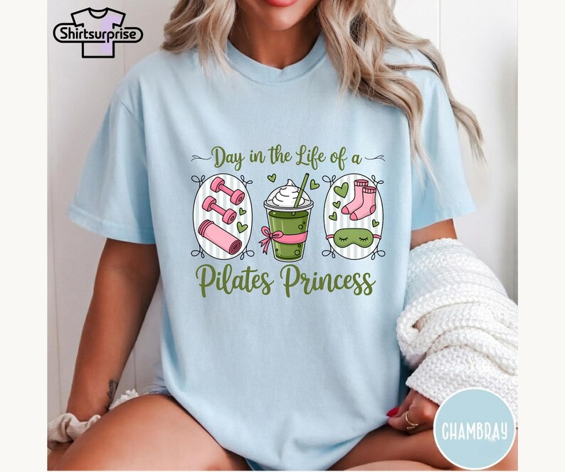 K&ouml;nnte beinhalten: Hellblaues T-Shirt mit dem Text "Day in the Life of a Pilates Princess". Die Grafik zeigt Hanteln, ein Getr&auml;nk, Socken und eine Schlafmaske. Das Shirt hat einen Rundhalsausschnitt und kurze &Auml;rmel.