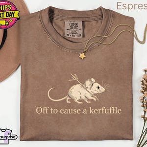 May include: Espresso-colored t-shirt with a graphic of a mouse holding a stick, and the text "Off to cause a kerfuffle." A gold star pendant necklace rests on the shirt. The shirt has a Comfort Colors tag.