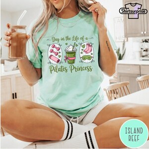K&ouml;nnte beinhalten: Hellgr&uuml;nes T-Shirt mit dem Text "Day in the Life of a Pilates Princess". Das Shirt zeigt Illustrationen von Hanteln, einem Getr&auml;nk und Socken. Das Shirt ist ein bequemer, l&auml;ssiger Stil.