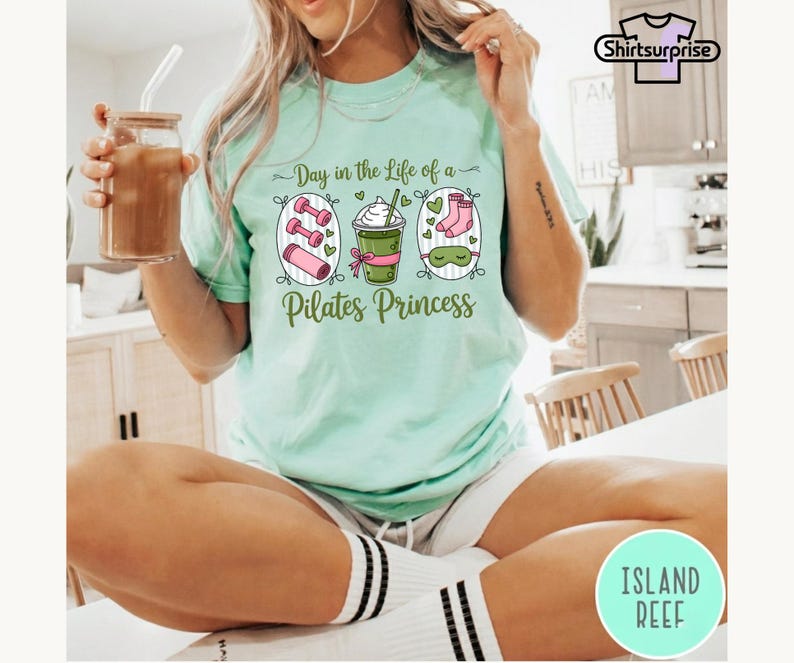 K&ouml;nnte beinhalten: Hellgr&uuml;nes T-Shirt mit dem Text "Day in the Life of a Pilates Princess". Das Shirt zeigt Illustrationen von Hanteln, einem Getr&auml;nk und Socken. Das Shirt ist ein bequemer, l&auml;ssiger Stil.