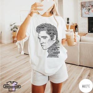 May include: A white short-sleeved t-shirt featuring a black graphic print of a portrait composed of text. The text includes song titles and phrases like My Way, Party, Jailhouse Rock, Teddy Bear, and It's Now or Never, forming the shape of a face.