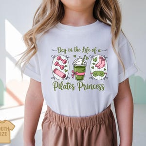 K&ouml;nnte beinhalten: Wei&szlig;es T-Shirt mit einem gr&uuml;n-rosa Aufdruck mit der Aufschrift "Day in the Life of a Pilates Princess". Die Grafik zeigt Hanteln, eine Yogamatte, ein Getr&auml;nk, Socken und eine Schlafmaske. Das Shirt ist mit "Youth Size" gekennzeichnet.