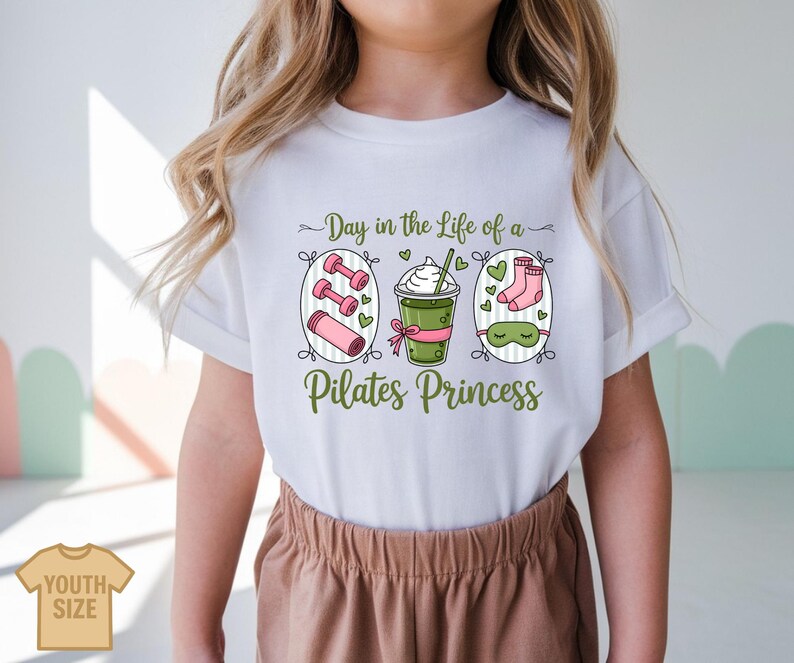 K&ouml;nnte beinhalten: Wei&szlig;es T-Shirt mit einem gr&uuml;n-rosa Aufdruck mit der Aufschrift "Day in the Life of a Pilates Princess". Die Grafik zeigt Hanteln, eine Yogamatte, ein Getr&auml;nk, Socken und eine Schlafmaske. Das Shirt ist mit "Youth Size" gekennzeichnet.