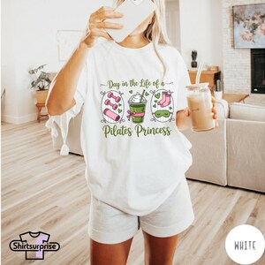 K&ouml;nnte beinhalten: Wei&szlig;es T-Shirt mit dem Text "Day in the Life of a Pilates Princess" und Illustrationen von Hanteln, einem Smoothie und Socken. Das Shirt wird mit grauen Shorts kombiniert. Eine Person h&auml;lt ein Telefon und ein Glas Eiskaffee.
