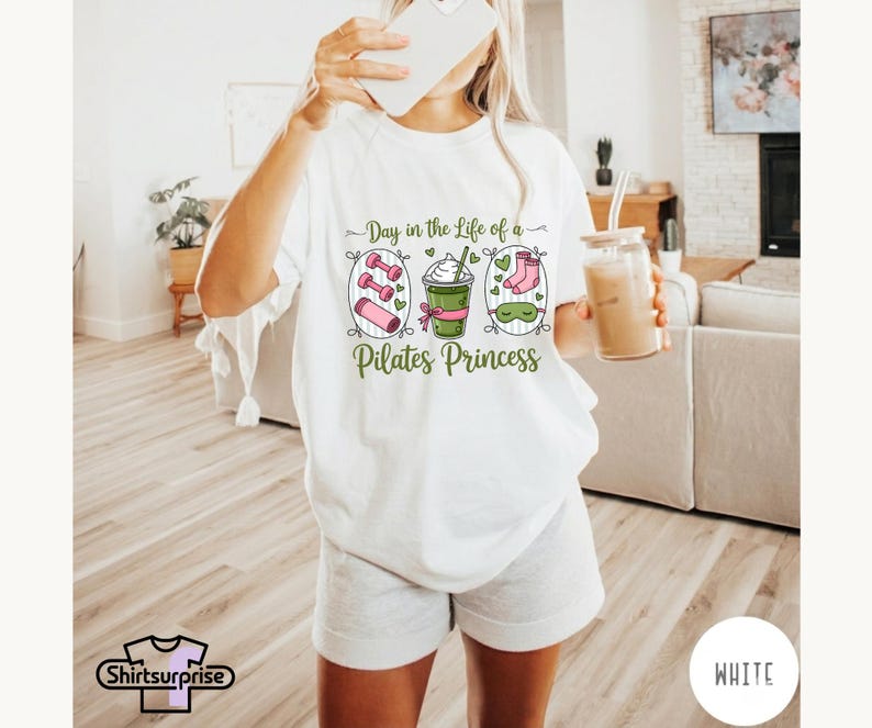 K&ouml;nnte beinhalten: Wei&szlig;es T-Shirt mit dem Text "Day in the Life of a Pilates Princess" und Illustrationen von Hanteln, einem Smoothie und Socken. Das Shirt wird mit grauen Shorts kombiniert. Eine Person h&auml;lt ein Telefon und ein Glas Eiskaffee.