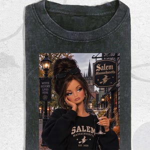 May include: A charcoal gray acid-wash t-shirt featuring a graphic print of a doll with an updo hairstyle and sunglasses, holding a martini glass. The print includes the text Salem Massachusetts, Witch History Lives Here, and Est. 1626.