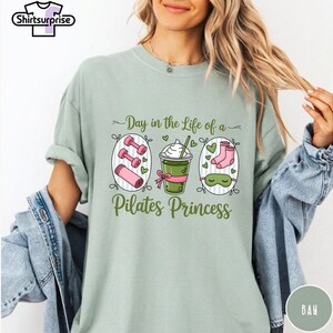 K&ouml;nnte beinhalten: Hellgr&uuml;nes T-Shirt mit dem Text "Day in the Life of a Pilates Princess". Das Shirt zeigt Illustrationen von rosa Hanteln, einem gr&uuml;nen Getr&auml;nk und einer Schlafmaske. Das Shirt hat einen bequemen, l&auml;ssigen Stil.