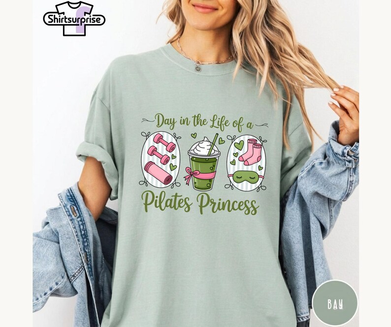 K&ouml;nnte beinhalten: Hellgr&uuml;nes T-Shirt mit dem Text "Day in the Life of a Pilates Princess". Das Shirt zeigt Illustrationen von rosa Hanteln, einem gr&uuml;nen Getr&auml;nk und einer Schlafmaske. Das Shirt hat einen bequemen, l&auml;ssigen Stil.