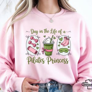 K&ouml;nnte beinhalten: Hellrosa Sweatshirt mit einem Grafikdesign, das Hanteln, eine Yogamatte, ein Getr&auml;nk, Socken und eine Schlafmaske zeigt. Der Text lautet: "Day in the Life of a Pilates Princess".