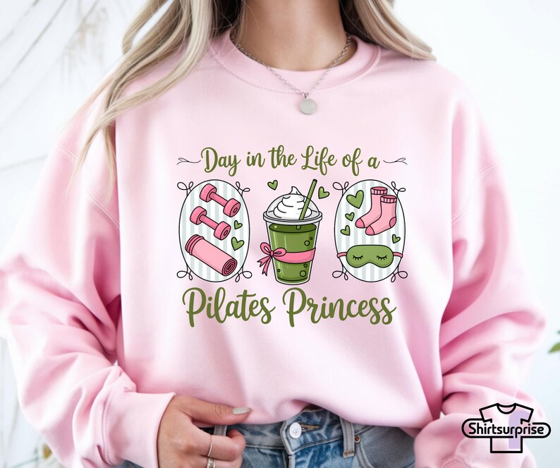 K&ouml;nnte beinhalten: Hellrosa Sweatshirt mit einem Grafikdesign, das Hanteln, eine Yogamatte, ein Getr&auml;nk, Socken und eine Schlafmaske zeigt. Der Text lautet: "Day in the Life of a Pilates Princess".