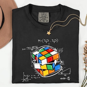 May include: Black t-shirt with a colorful Rubik's Cube graphic and mathematical equations. A gold star pendant necklace rests on the shirt. The shirt has a Comfort Colors tag, size XL-TO-EO.