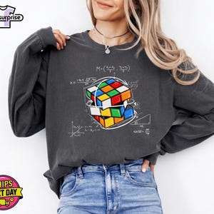 May include: Dark gray sweatshirt featuring a colorful Rubik's Cube graphic with mathematical equations. The design includes a multi-colored cube with red, blue, green, yellow, orange, and white squares. The sweatshirt has long sleeves and a crew neck.