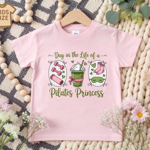 K&ouml;nnte beinhalten: Hellrosa Kinder-T-Shirt mit dem Text "Day in the Life of a Pilates Princess". Die Grafik zeigt Hanteln, ein Getr&auml;nk, Socken und eine Schlafmaske. Das T-Shirt h&auml;ngt an einem Holzkleiderb&uuml;gel.