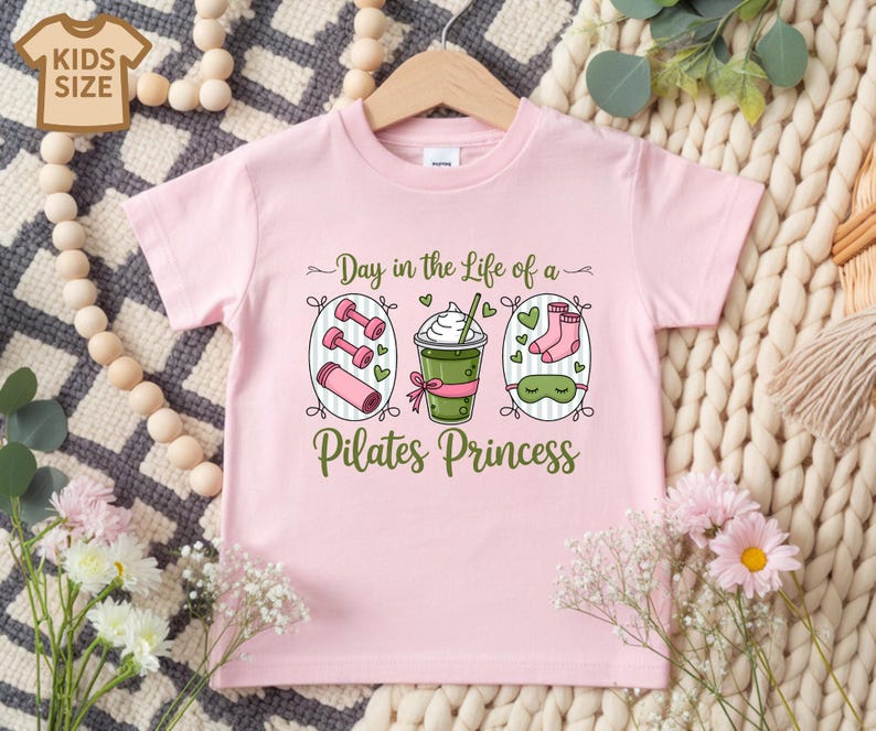 K&ouml;nnte beinhalten: Hellrosa Kinder-T-Shirt mit dem Text "Day in the Life of a Pilates Princess". Die Grafik zeigt Hanteln, ein Getr&auml;nk, Socken und eine Schlafmaske. Das T-Shirt h&auml;ngt an einem Holzkleiderb&uuml;gel.