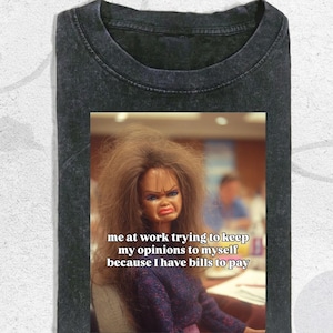 May include: A charcoal grey acid-wash t-shirt featuring a graphic of a doll with messy brown hair and a frustrated facial expression. The text on the graphic reads: me at work trying to keep my opinions to myself because I have bills to pay.