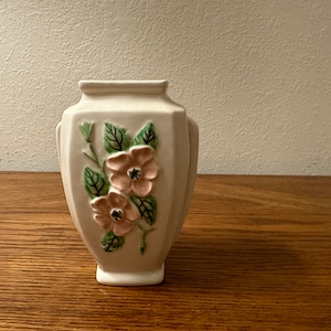 May include: Cream-coloured ceramic vase with a floral pattern. The vase has two pink flowers with green leaves and stems. It has a rectangular shape with curved sides and two small handles.
