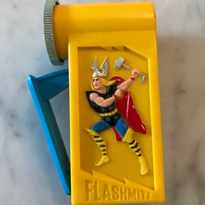 May include: Yellow plastic flashlight with a blue handle. The front of the flashlight features a cartoon image of Thor from Marvel Comics, wielding his hammer. The text "FLASHMITE" is printed on the bottom of the flashlight.