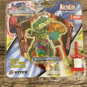 May include: A Transformers Energon brand Microlite Ylar kite with a 1-foot wingspan. The kite features a green and yellow robot character with a red, white, and blue tail. The packaging includes a red and white string and a red handle.