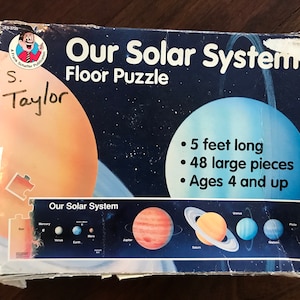 May include: A blue and orange box for a floor puzzle of our solar system. The box shows a depiction of the planets and the sun. The box is 5 feet long and has 48 large pieces. It is recommended for ages 4 and up.