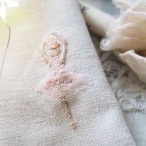 May include: A delicate pink embroidered ballerina figure on a white linen fabric background.