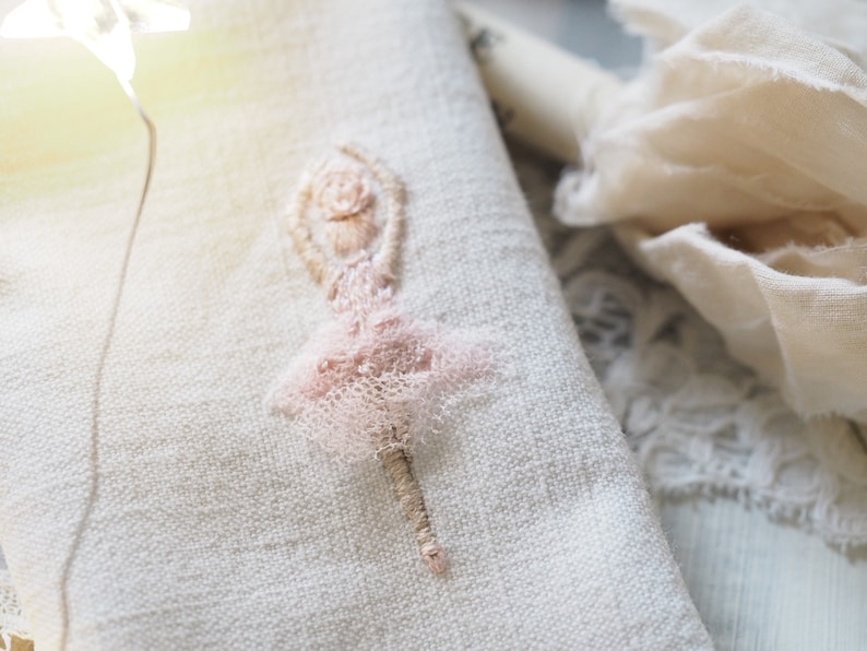May include: A delicate pink embroidered ballerina figure on a white linen fabric background.