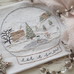 Może przedstawiać: Białe płótno haftowane wzorem kuli śnieżnej przedstawiającej mały domek, drzewa, bałwana i postać jeżdżącą na łyżwach. Kula śnieżna jest otoczona brązową ramką z napisem "let it snow" napisanym kursywą.