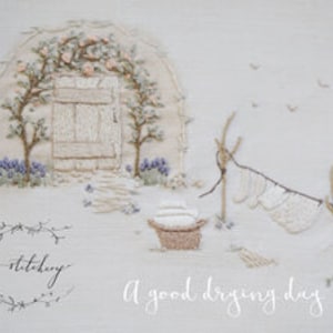 Könnte beinhalten: Eine weiße Stickerei mit einem Häuschen mit einer Tür, zwei Bäumen und einer Wäscheleine mit hängender Wäsche. Der Text "A good drying day" befindet sich unter dem Bild.