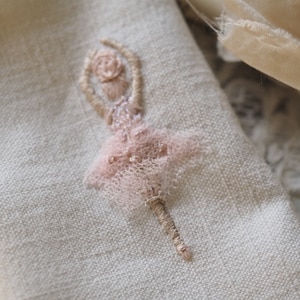 May include: A delicate pink embroidered ballerina on a white linen fabric.