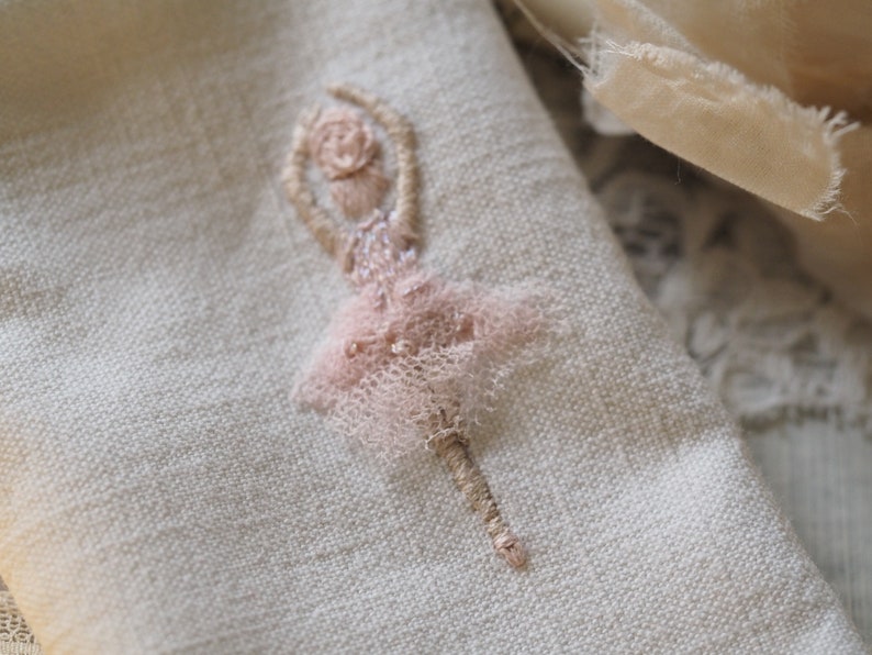May include: A delicate pink embroidered ballerina on a white linen fabric.
