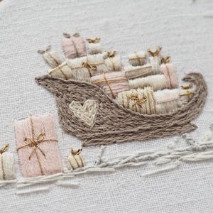 Może przedstawiać: Sanie wyszywane w kolorze brązowym i białym, wypełnione różowymi i białymi zapakowanymi prezentami. Sanie mają kształt serca z boku. Sanie znajdują się na białym tle z wyszywaną szarą ścieżką z tyłu.