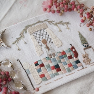 May include: A white fabric with a hand-stitched embroidery design of a girl in a bed with a patchwork quilt. The embroidery includes a canopy over the bed, a small tree, and a small stuffed animal.