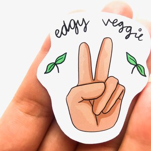 May include: White sticker with a peace sign hand gesture and the text "edgy veggie" in black cursive. Two green plant sprouts are on either side of the peace sign.