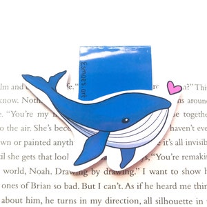 Op de afbeelding: Een blauwe walvis-bladwijzer met een witte buik en een roze hart. De bladwijzer is gemaakt van papier en heeft een glanzende afwerking. De tekst "Esmaes.art" is op de bladwijzer gedrukt.