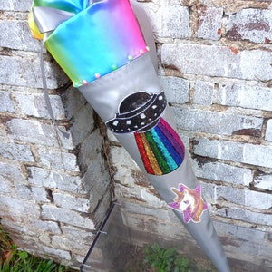 May include: A gray, cone-shaped school bag with a rainbow satin top and a large bow. The bag features a beaded UFO design with a rainbow beam and a unicorn patch. The bag is leaning against a brick wall.
