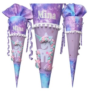 Può includere: Tre contenitori regalo a forma di cono con tessuto a stampa galassia viola e blu sulla parte superiore e inferiore. Ogni cono mostra il nome Mina in lettere bianche, un'applicazione unicorno bianco con criniera in lana multicolore e pompon bianchi.