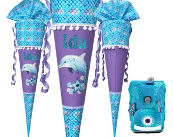 Puede incluir: Dos conos escolares de color morado y azul con un diseño de delfín y el nombre "Ida" en purpurina. También se incluye una mochila azul a juego con un diseño de delfín.