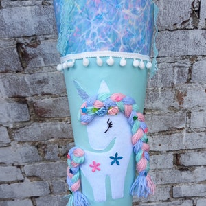 Può includere: Un cono scolastico a forma di unicorno blu chiaro con un viso di unicorno bianco, trecce di lana rosa e blu e un bordo in pizzo blu chiaro e bianco.
