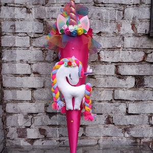 May include: A pink unicorn school cone with a white unicorn, rainbow mane, and a gold horn. The cone is decorated with flowers and glitter.