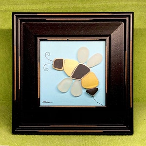 May include: A framed artwork featuring a bee made from sea glass and painted on a blue background. The bee is yellow, black, and white.