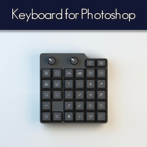 Könnte beinhalten: Eine schwarze Tastatur mit weißen Symbolen für Photoshop. Die Tastatur hat zwei Knöpfe oben und eine Vielzahl von Tasten mit Symbolen für verschiedene Photoshop-Tools.