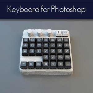 Può includere: Una tastiera personalizzata per Photoshop con tasti neri e icone bianche. La tastiera ha una base strutturata bianco sporco, quattro manopole cilindriche e la scritta "Keyboard for Photoshop" in alto.