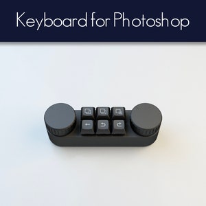 Könnte beinhalten: Eine schwarze Tastatur, die für die Verwendung mit Photoshop entwickelt wurde. Sie verfügt über eine Reihe von vier Tasten mit Symbolen für verschiedene Photoshop-Tools und eine Reihe von drei Tasten mit Pfeilen für die Navigation. Die Tastatur hat außerdem zwei runde Knöpfe an beiden Seiten.