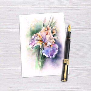 Peut inclure: Une peinture à l'aquarelle d'une fleur d'iris jaune et violette sur une carte blanche. La carte est à côté d'un stylo plume noir et or.