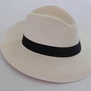 May include: A cream-colored woven straw fedora hat featuring a pinched crown and a wide brim. The hat is accented with a solid black grosgrain ribbon band encircling the base of the crown, providing a distinct contrast to the light-colored straw material.