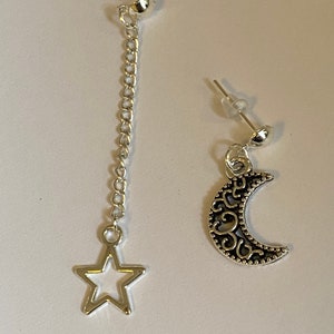 May include: A pair of silver earrings, one with a dangling star charm on a chain and the other with a crescent moon charm.