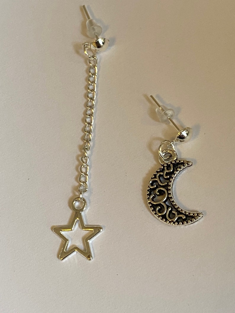 May include: A pair of silver earrings, one with a dangling star charm on a chain and the other with a crescent moon charm.