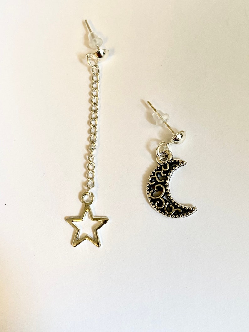 May include: A pair of silver earrings with a dangling star and crescent moon charm. The earrings are on a white background.
